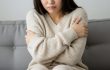 寒さに耐える若い日本人女性のイメージ。クリーム色のニットセーターを着て、腕を抱えるようにして寒そうな仕草をしている。顔は一部が見えるだけで、寒さによる不快感が表情から伝わる。背景はソファとぼんやりとしたニュートラルな色合いで、冷え性の雰囲気を強調している。